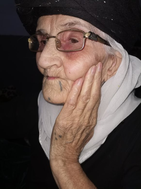 A photo of Nemsha Shlo Smo. They are wearing black clothing and a white scarf. She has a black tattoo dot and line on her chin, and three black dots in a triangle shape on her hand. Credit: Nidaa Khalil Aswad 