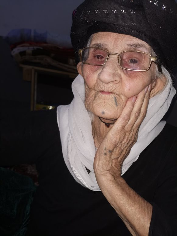 A photo of Nemsha Shlo Smo. They are wearing black clothing and a white scarf. She has a black tattoo dot on her chin. Credit: Nidaa Khalil Aswad 