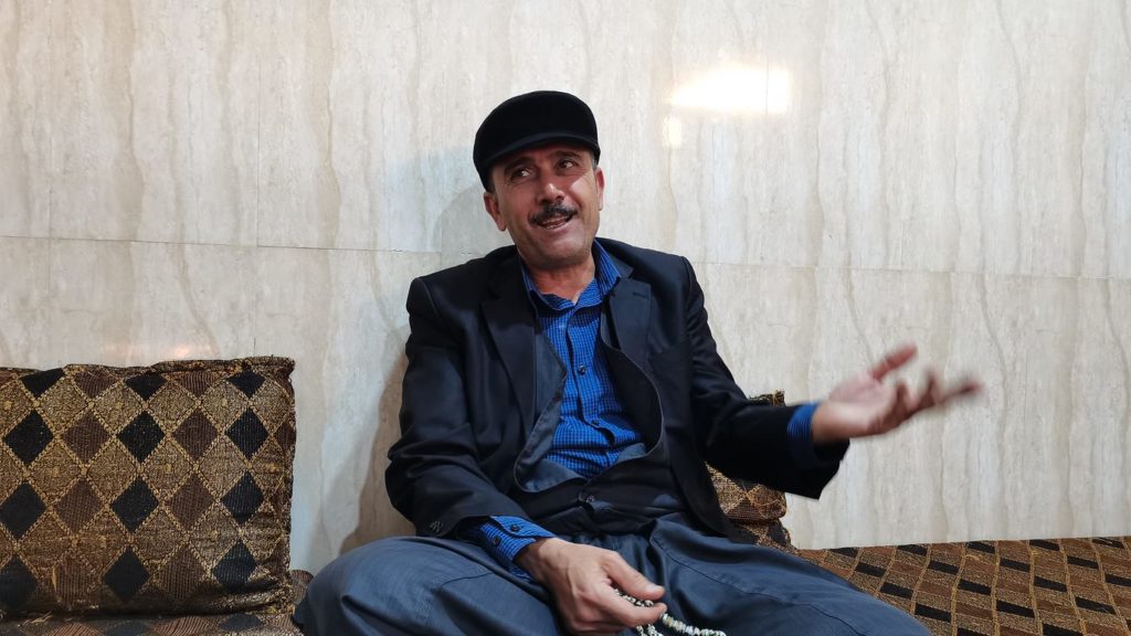 A photo of Raad Eido Rehan. He is wearing a black jacket and hat, with a blue shirt. He rests against a large marble wall with brown cushions. Credit:  Irvin Ibrahim Solman 