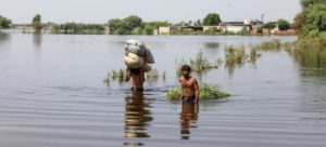 Two men walking in waist-high floodwater in Pakistan, one carrying two large bags on his shoulder
